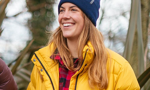 Smiling woman wearing a navy blue beanie and a yellow puffy down jacket in the woods