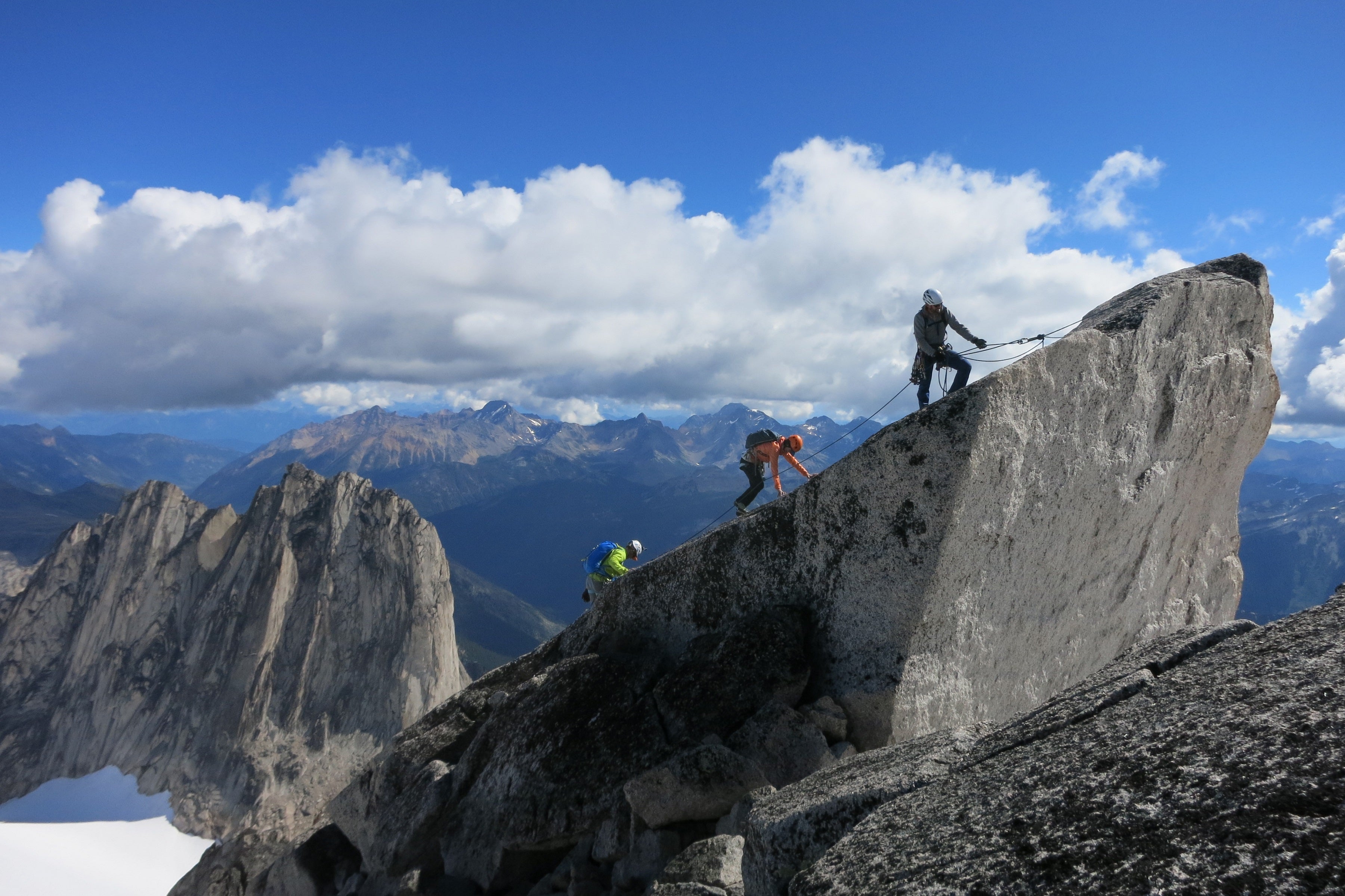One climber top belays two others near the peak of a mountain. Mountains are visible in the background.